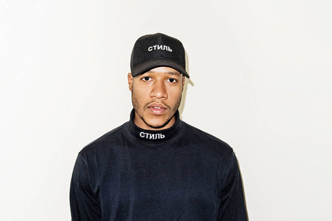 An image of a Black man against a white-grey background wearing a black mockneck shirt and a black baseball hat.