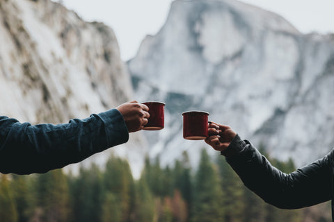 An image of two outstretched hands cheers'ing with coffee cups, with mountains in the background.