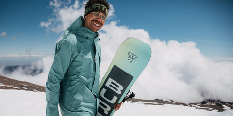 An image showing snowboarder Kody Williams holding a snowboard, in the background is a snow-covered hill and a cloudy blue sky.