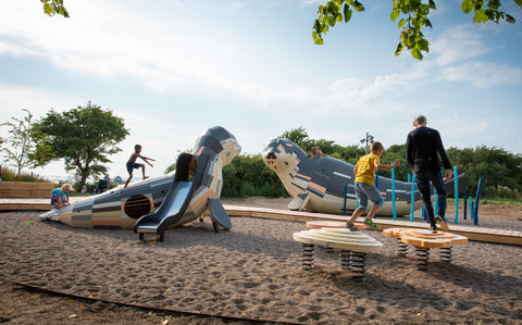 An image of a playground featuring sculptures of sea mammals with kids running around on the sand.