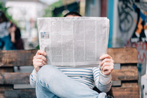 A picture of a person seated on a bench reading a newspaper, the newspaper obscuring their face and torso.