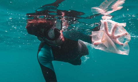 A person swimming in scuba gear, reaching a hand out to collect a plastic bag.