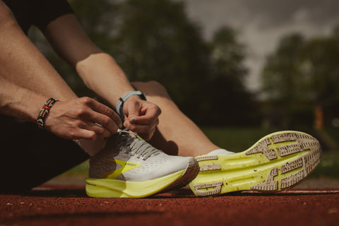 An image of a person sitting on an outdoor sports track, lacing up the shoelaces of their left shoe, shown from the knees down.