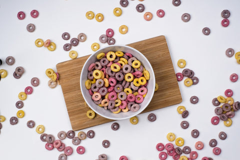 An overhead image of a bowl of cereal on a wooden cutting board. Cereal fills the bowl and is spread around the cutting board.
