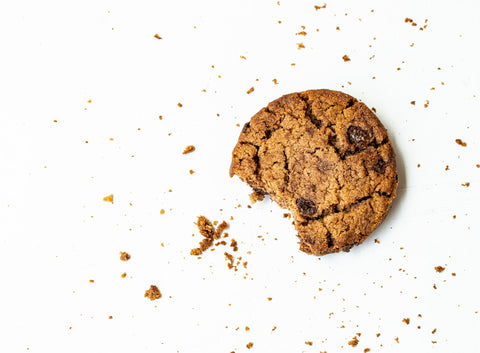 An image of a cookie on a white background. The cookie has a bite taken out of it and crumbs lay in the space between the bite and the white surface.