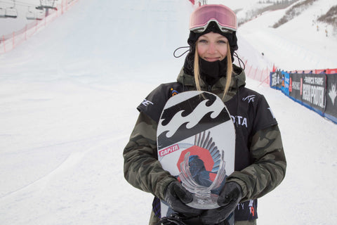 An image of Jasmine Baird holding her snowboard to her chest while wearing a ski jacket and big ski goggles, standing at the base of a ski hill.