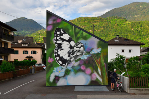 Another image of a different photo-realistic butterfly perched on a flower on another building. In the background are rolling green hills and smaller buildings.