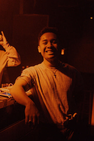 An image of Steven Gamboa with an orange-yellow light overhead. He relaxes with his arm on a table while wearing a white t-shirt and a big smile.