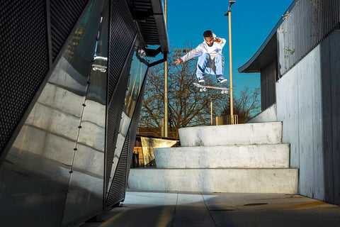 An image of Malik Walker, a young Black man, leaping through the air on his skateboard as he cruises over concrete stairs.