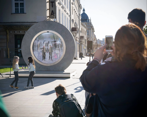 An image of a crowd of people stood in front of a life-size round portal-like screen, taking photos as people wave from inside the object.