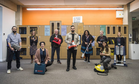 An image of a group of people standing in front of tan-colored lockers, each of whom is holding an object like a sewing machine or vacuum cleaner.