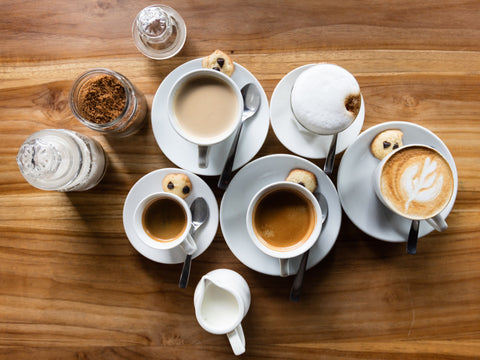 A messy-looking collection of coffee and tea cups on a wooden surface with a cream and sugar bowl added to the mix.