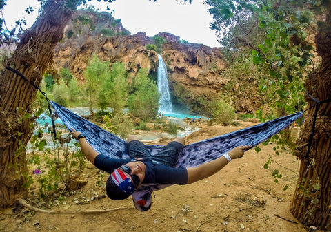 Young man leaning back in his hammock, hung between two trees in front of a waterfall