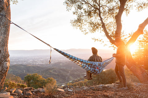 Woman wearing carhartt beanie sits in hammock during sunset overlooking a cityscape with man in puffer coat