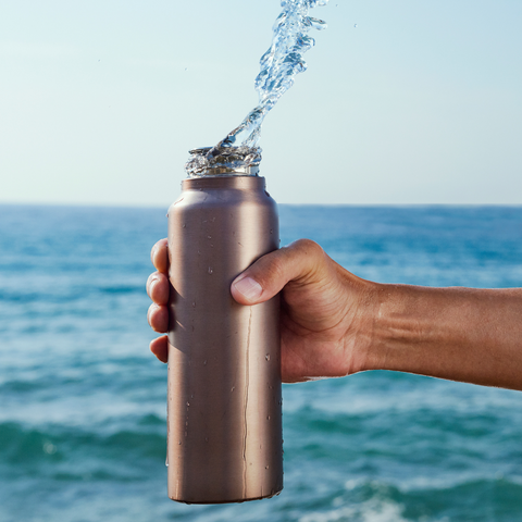 A person holding a tin reusable water bottle with water splashing upwards.
