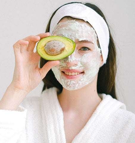 Stock photo of a woman holding an avocado with a face mask on.
