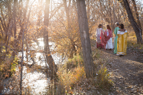 four women dressed in corsets and flowing robes walk near a stream in late afternoon autumn light