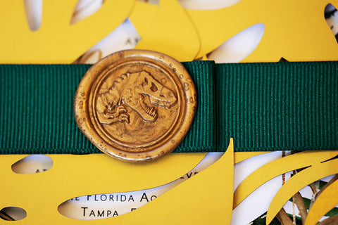 Picture of a wedding invitations wrapped in a yellow leaf cut out form paper, tied with a green ribbon and sealed with golden wax pressed with the Jurassic Park Logo. 