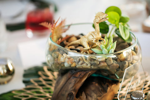 Picture of a wedding table center place that looks like a terrarium featuring a little golden dinosaur figurine to fit the Jurassic Park Wedding Theme.  