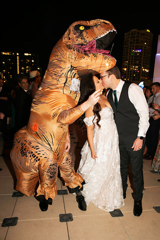 Picture of a bride and groom kissing next to a person wearing an inflatable dinosaur costume