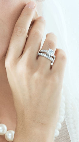 Bride wearing a bridal ring set on the ring finger of her left hand. The engagement ring has a large round cut diamond and the band is inset with smaller diamonds. Her wedding band is an eternity band design, a ring inset with diamond of a uniform size all the way around the band.