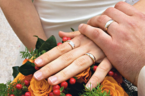 Bride and groom lay their left hands over the bride's bouquet of orange and red flowers. Bride is wearing a simple white gold wedding band on her left hand's ring finger.  Bride is wearing another ring on her pointer finger that has two diamonds in a Toi Et Moi design. Groom is wearing a simple white gold band on the ring finger of his left hand.