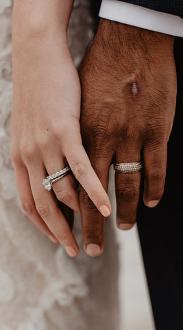 A bride and groom's left hands posed together by their sides. The bride's rings are an white gold engagement ring with a large pronged diamond in the center and her wedding band is a thick eternity band with uniform sized diamonds encircling the exterior of the band. The groom's band is yellow gold and encrusted with small diamonds.