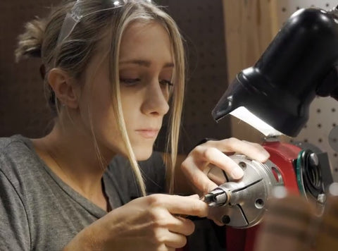 Kassie, a ring crafter, looks down at the ring she is crafting on her lathe, using both hands to craft with a lamp shining in to the right of the frame down on the lathe Kassie is working at. 