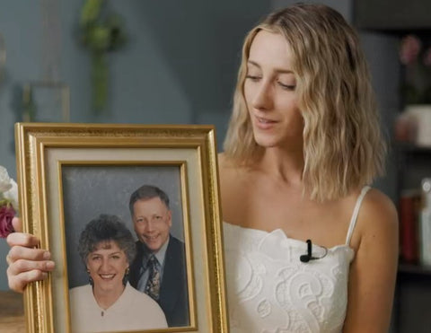Blonde woman wearing white dress and black mic, holding a golden frame with a picture of an older couple, a woman with dark hair, wearing a pale blouse, and a grey-haired man, wearing a suit