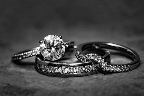 Three rings posed together. The left is a white gold engagement ring with a large round cut diamond with a pronged setting. The middle ring is a eternity band with uniform diamonds inset along the exterior of the white gold band. The right ring is a second wedding band inset with small diamonds that has a notched feature.