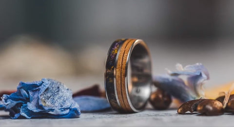 Light blue, purple, and yellow flower petals scattered around a titanium band inlayed with flower petals of purple and yellow, coffee grounds, and white oak wood