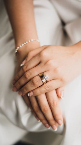 A bride sits posed with her left hand on top of her right hand. On her left hand's ring finger she is wearing her wedding band atop her engagement ring. The engagement ring has a large round stone. Her wedding band is thick yellow gold with a small inset diamond.