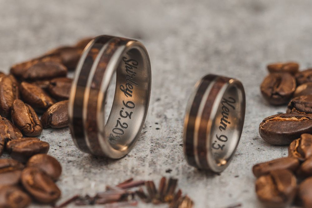two-titanium-rings-inlaid-with-one-channel-of-columbian-coffee-grounds-and-one-channel-of-rutile-and-engraved-inside-both-bands-are-each-spouses-first-name-and-their-wedding-date-the-ring-sit-on-cool-light-grey-concrete-and-are-surrounded-by-whole-roasted-coffee-beans