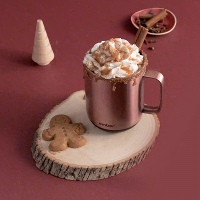 A gif of the Rose Gold Ember Mug² filled with whipped cream and cinnamon sits on a round cut of wood along wih two gingerbread cookies. The background is a deep red and is filled with gingerbread trees and cinnamon sticks. A hand places another gingerbread cookie.