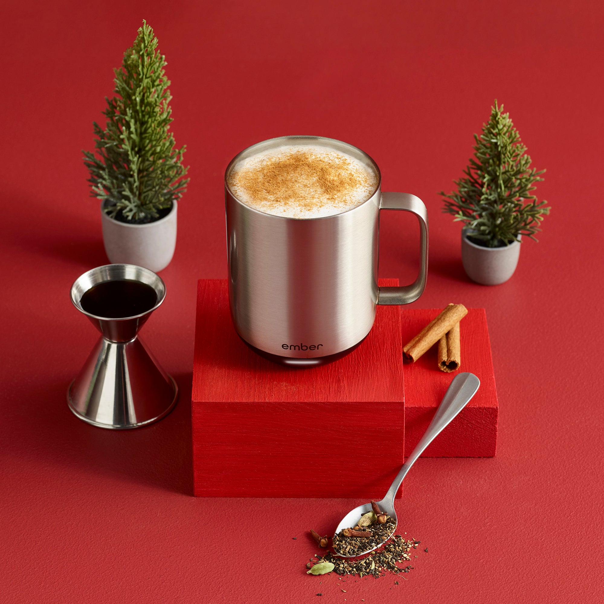A Stainless Steel Ember Mug² filled with steamed milk and cinnamon sits atop a a red wooden block. It is surrounded by small leafy trees, cinnamon sticls, a spoonful of black tea, and a stainless steel jigger of syrup. The background is a festive red color.