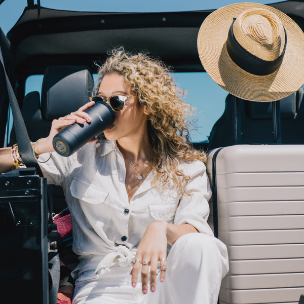 Womam sits in the back of the Jeep, suitcase on her right, sipping from the Ember Travel Mug.