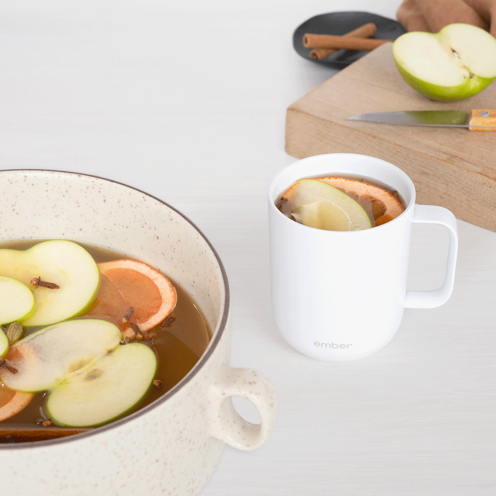 A white ember mug filled with apple cider and topped with apple slices next to a cutting board, a bowl with various spices, and a pot of mulled apple cider with the apples and oranges swirling.