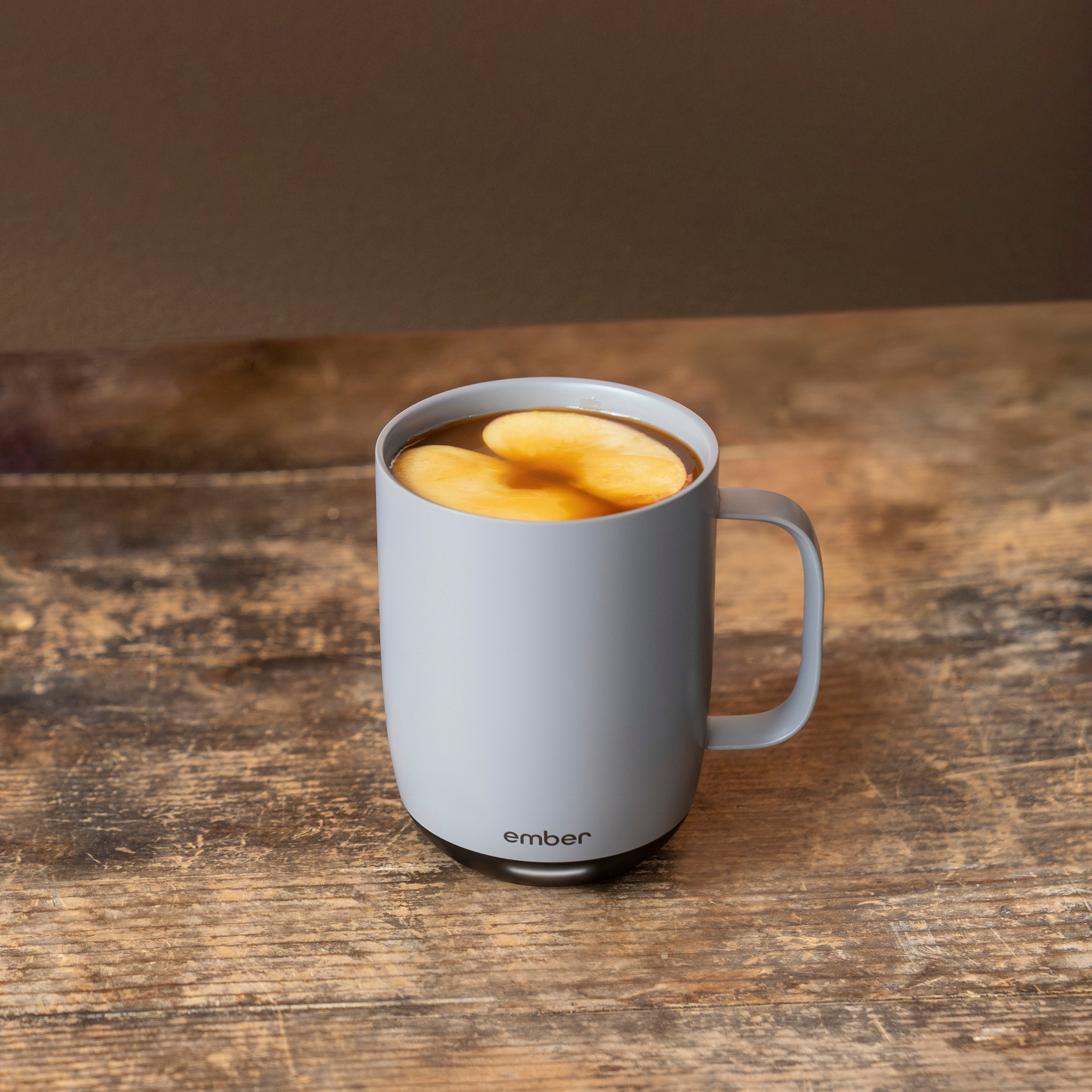 A Gray Ember Mug² is filled with a hot toddy drink and topped with apple slices. It sits on a weathered dark wooden table.