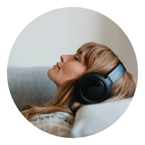 While laying down on a couch with her eyes closed, a young woman listens to music coming out of her chunky black headphones.