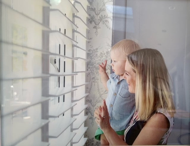 mum with a little child in her arms looking out a window with shutters