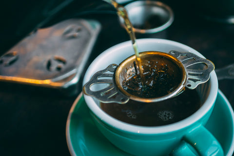 A person pouring loose leaf Earl Grey tea into a blue mug through a strainer