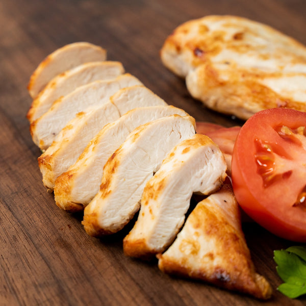 Close-up of a sliced, cooked Poulet Rouge® Heritage Chicken boneless skinless breast on a wooden cutting board. The juicy, golden-brown slices showcase the tender, flavorful meat, accompanied by a fresh tomato slice, highlighting the superior quality of Joyce Farms' slow-growing chicken."