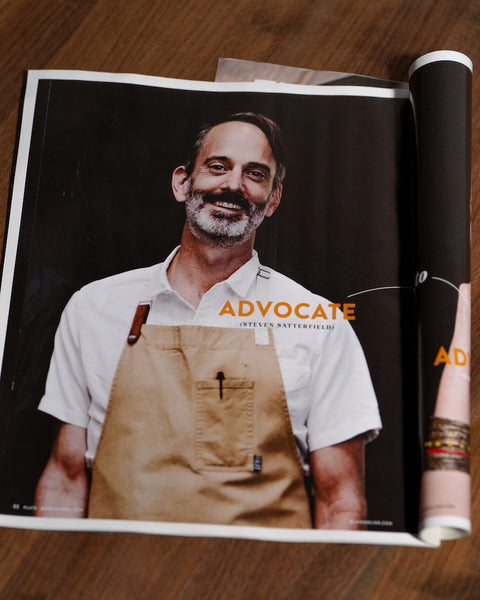 This image shows an open magazine featuring a photo of Chef Steven Satterfield from Miller Union Restaurant. The chef, wearing a crisp white shirt and a tan apron, is smiling and appears relaxed. The title "Advocate" is prominently displayed next to him, suggesting a feature story within the magazine. The magazine is resting on a wooden surface, with its pages slightly rolled up towards the binding, indicating it's been well-read or flipped through.