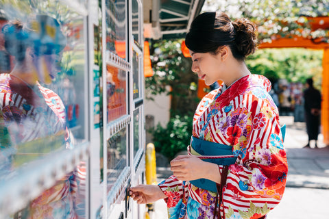 Japanese girl wearing kimono buying cold drinks from the vending machine. Kyoto woman throwing coins and choosing a snack or drink at vending machine on sunny day.