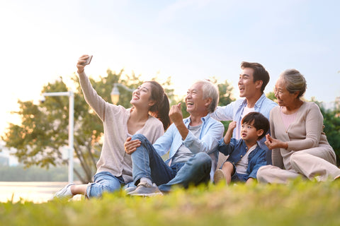 A three-generation family is sitting in the grass at a park and taking a picture using a cell phone