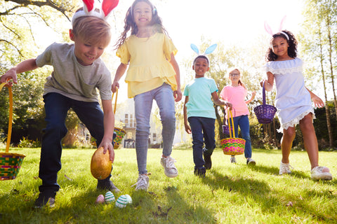 Group Of Children Wearing Bunny Ears Running To Pick Up Chocolate Egg On Easter Egg Hunt In Garden