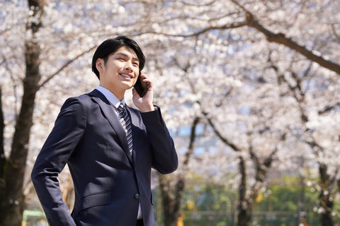 A Japanese businessman making a phone call to employers with cherry blossoms in the background.