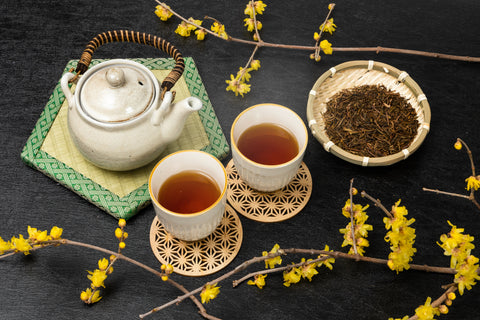 A teapot along with two tea cups filled with hojicha tea and a bowl of hojicha leaves.