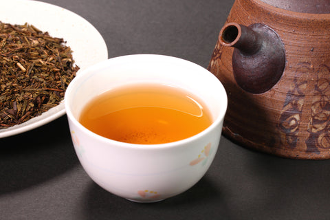 A tea cup filled with hojicha roasted green tea alongside a teapot and plate of tea leaves.