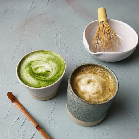 A matcha latte, left, and a hojicha latte, right, on a table along with a bowl and whisk.